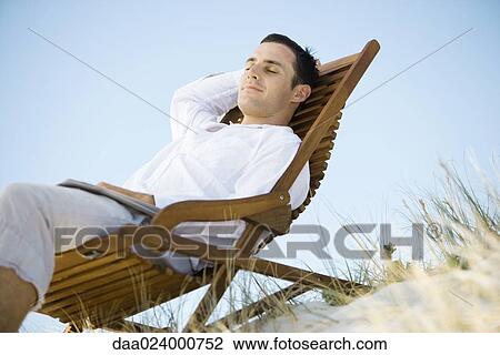 Young man sitting in deck chair, smiling, low angle view View Large Photo Image Stock Image - Young man sitting in deck chair, smiling, low angle view. Fotosearch