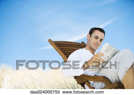 Young man sitting in deck chair, reading newspaper View Large Photo Image Stock Photography - Young man sitting in deck chair, reading newspaper. Fotosearch