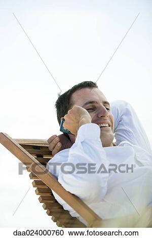 Stock Image - Young man sitting in deck chair, using cell phone. Fotosearch
