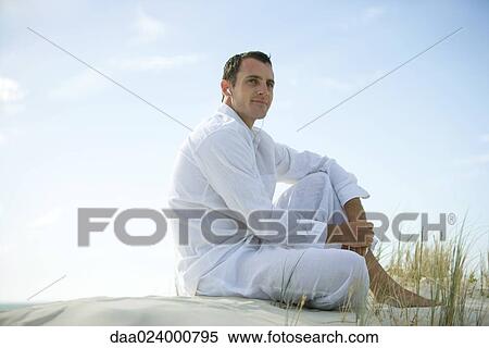 Young man sitting on dune, listening to earphones, smiling, looking away View Large Photo Image Stock Photography - Young man sitting on dune, listening to earphones, smiling, looking away. Fotosearch