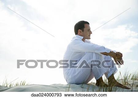 Young man sitting on dune, smiling, full length View Large Photo Image Stock Image - Young man sitting on dune, smiling, full length. Fotosearch