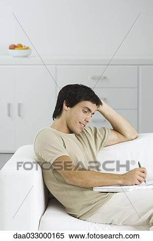 Young man sitting on sofa, using laptop computer as hard surface to write on View Large Photo Image Stock Photography - Young man sitting on sofa, using laptop computer as hard surface to write on. Fotosearch
