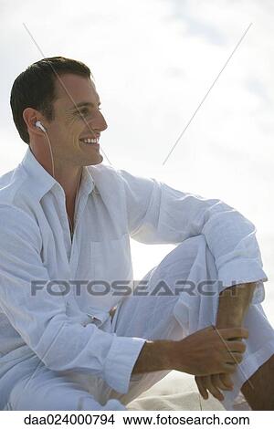 Picture - Young man sitting with knee up, listening to earphones, smiling, looking away. Fotosearch