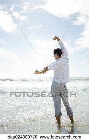 Young man standing in surf, stretching, rear view View Large Photo Image Stock Photo - Young man standing in surf, stretching, rear view. Fotosearch