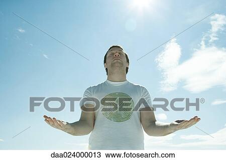 Stock Image - Young man standing in the sun with eyes closed and arms out. Fotosearch