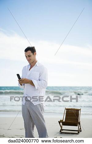 Stock Image - Young man using cell phone on beach. Fotosearch