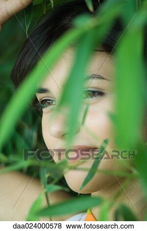 Stock Photo - Young woman among foliage, smiling, looking away, close-up. Fotosearch