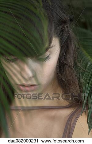 Stock Image - Young woman behind palm frond, looking down sadly. Fotosearch
