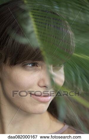 Stock Photo - Young woman behind palm frond, looking away in thought. Fotosearch