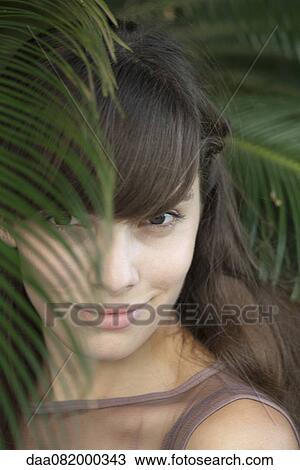Stock Image - Young woman behind palm frond, portrait. Fotosearch
