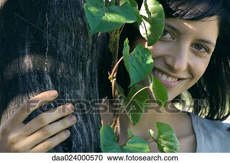 Young woman behind tree trunk, smiling at camera, portrait View Large Photo Image Stock Image - Young woman behind tree trunk, smiling at camera, portrait. Fotosearch