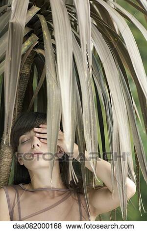 Stock Photo - Young woman behind tropical foliage, hand covering one eye. Fotosearch