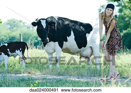 Stock Image - Young woman bending over in front of cows, smiling at camera. Fotosearch