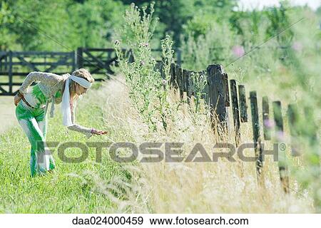 Stock Photo - Young woman bending over to touch tall grass, side view. Fotosearch