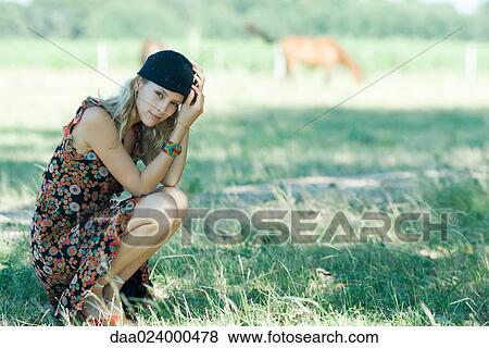 Stock Photo - Young woman crouching in field, smiling at camera. Fotosearch