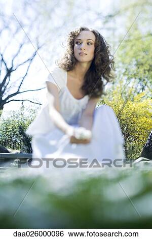 Stock Photograph - Young woman crouching outdoors, looking away, viewed from surface level. Fotosearch