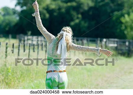 Stock Photo - Young woman dressed as hippy standing in field with arms out, rear view. Fotosearch