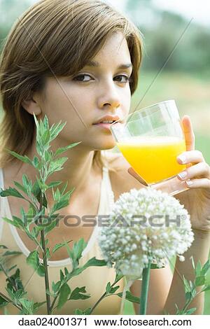 Stock Image - Young woman drinking glass of orange juice, plants in foreground, close-up. Fotosearch