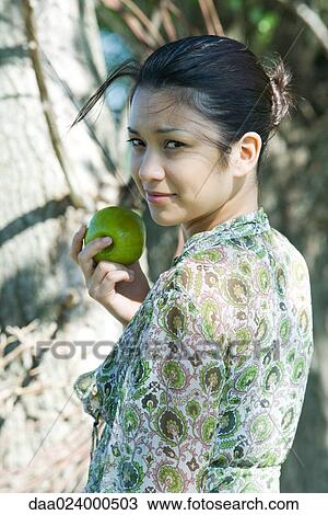 Stock Image - Young woman holding apple and looking at camera over her shoulder. Fotosearch