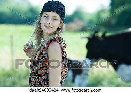 Young woman holding flower in rural setting, smiling at camera, cows in background View Large Photo Image Picture - Young woman holding flower in rural setting, smiling at camera, cows in background. Fotosearch