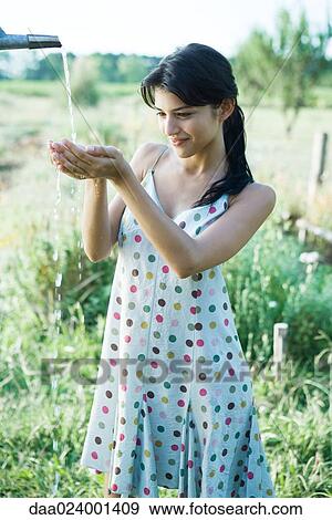 Stock Photo - Young woman holding hands under outdoor faucet, three quarter length. Fotosearch