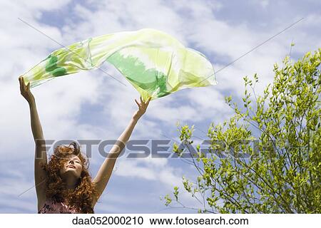 Stock Image - Young woman holding scarf up in breeze. Fotosearch