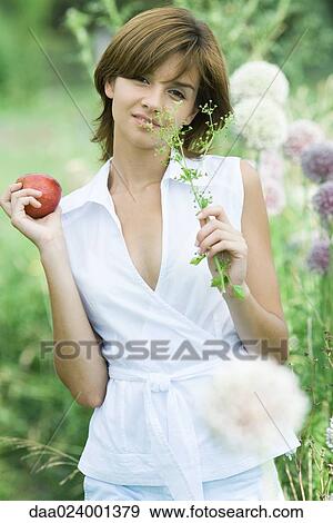 Stock Photo - Young woman holding sprig of herbs and apple, looking at camera. Fotosearch