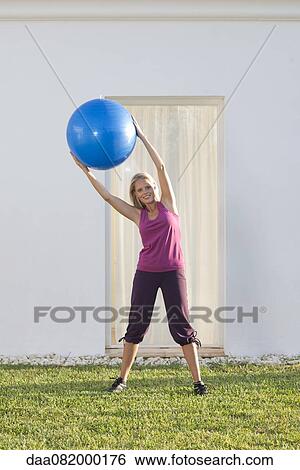 Stock Photograph - Young woman holding up fitness ball, leaning to one side. Fotosearch