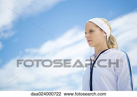 Stock Image - Young woman in active wear, listening to earphones, low angle view. Fotosearch