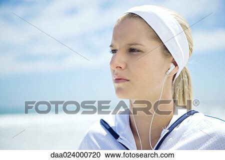 Stock Image - Young woman in active wear on beach, listening to earphones. Fotosearch