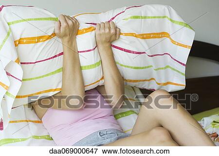 Stock Photo - Young woman in bed, covering face with pillow. Fotosearch