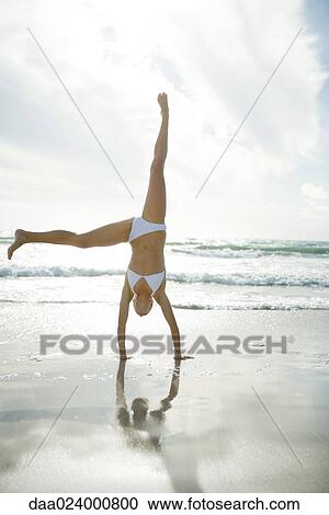Stock Image - Young woman in bikini doing cartwheel on beach, full length. Fotosearch