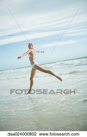 Stock Image - Young woman in bikini, leaping in surf, in mid air. Fotosearch
