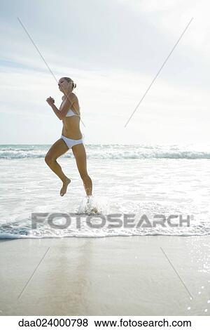 Stock Photo - Young woman in bikini, running and jumping in surf. Fotosearch