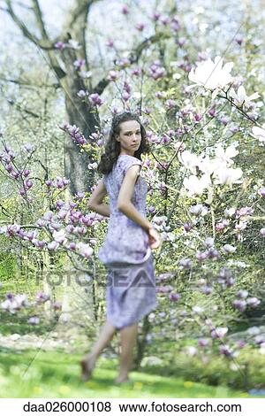 Stock Photo - Young woman in dress walking beside flowering tree, looking over shoulder at camera. Fotosearch