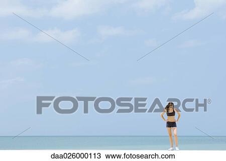 Young woman in sports clothing standing at the beach, looking down, mid-distance View Large Photo Image Stock Image - Young woman in sports clothing standing at the beach, looking down, mid-distance. Fotosearch