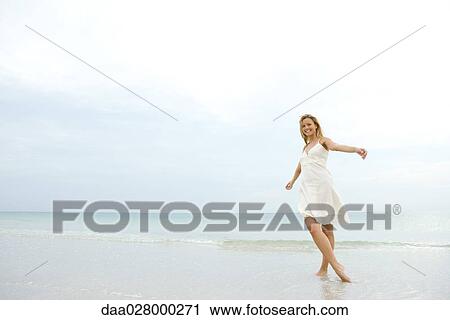 Stock Image - Young woman in sundress posing on beach with arms out and legs crossed, smiling at camera. Fotosearch