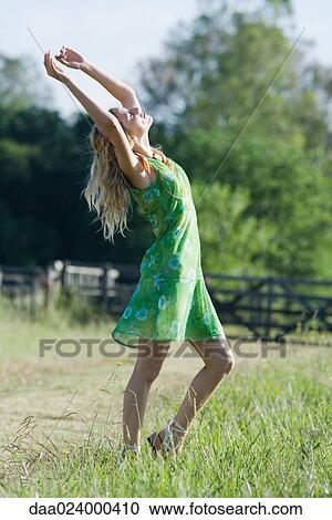 Stock Image - Young woman in sundress standing in rural field with arms raised. Fotosearch