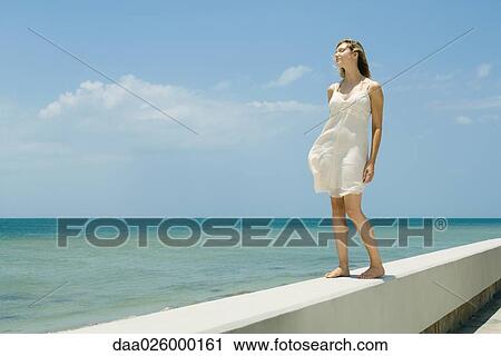 Stock Image - Young woman in sundress standing on low wall beside sea, eyes closed. Fotosearch