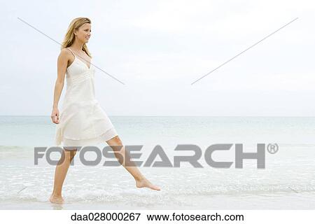 Young woman in sundress taking large step as she walks in shallow water on beach, full length View Large Photo Image Stock Photo - Young woman in sundress taking large step as she walks in shallow water on beach, full length. Fotosearch