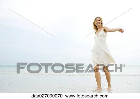 Stock Image - Young woman in sundress walking in surf, arms out, looking away. Fotosearch