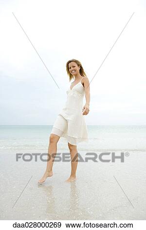 Young woman in sundress walking on wet sand at beach, smiling at camera, full length View Large Photo Image Stock Photo - Young woman in sundress walking on wet sand at beach, smiling at camera, full length. Fotosearch