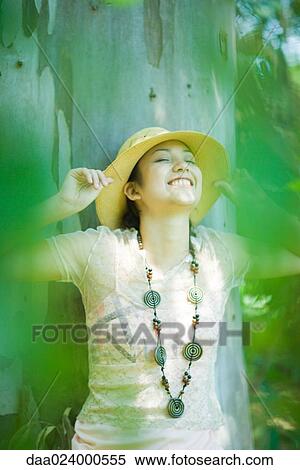 Young woman in sunhat leaning against tree trunk, smiling, eyes closed View Large Photo Image Stock Photography - Young woman in sunhat leaning against tree trunk, smiling, eyes closed. Fotosearch
