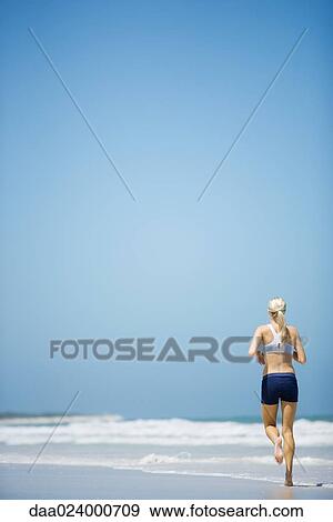 Stock Photo - Young woman jogging on beach, rear view, full length. Fotosearch