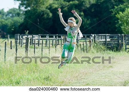 Stock Image - Young woman jumping in rural field with arms raised. Fotosearch
