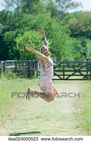 Stock Image - Young woman jumping with arms raised in rural setting, smiling at camera. Fotosearch
