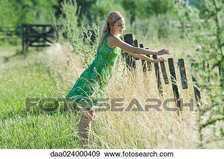 Stock Photo - Young woman leaning against fence in rural field, smiling. Fotosearch