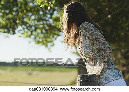 Picture - Young woman leaning against fence, looking into distance. Fotosearch