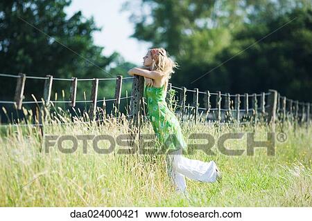 Stock Image - Young woman leaning against rural fence with eyes closed. Fotosearch