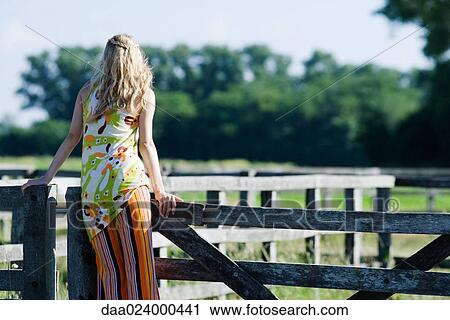 Stock Image - Young woman leaning against rural fence, rear view. Fotosearch
